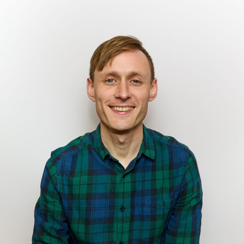 Smiling man in a green and blue plaid shirt against a light plain background.