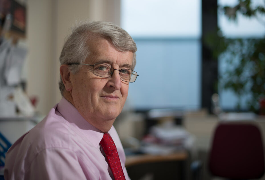 Portrait of an older man with gray hair and glasses, wearing a pink shirt and red tie in an office setting
