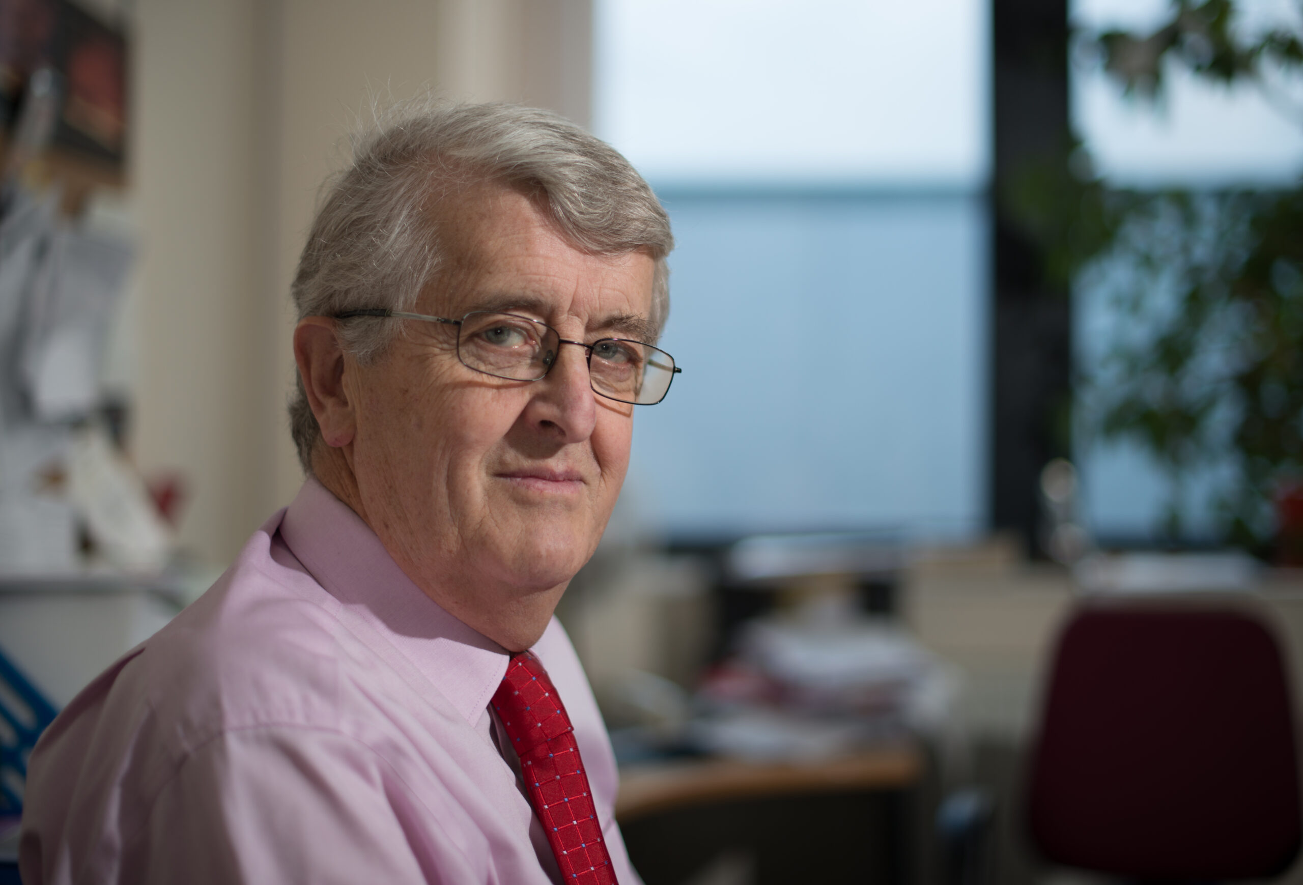 Portrait of an older man with gray hair and glasses, wearing a pink shirt and red tie in an office setting