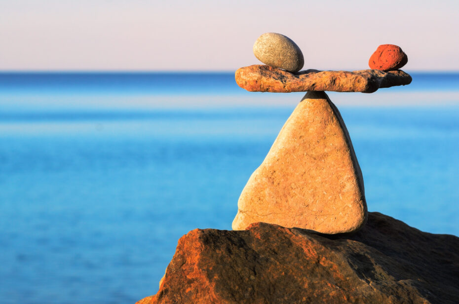 Stacked rocks balanced on a large rock by the blue ocean, with calm water in the background.