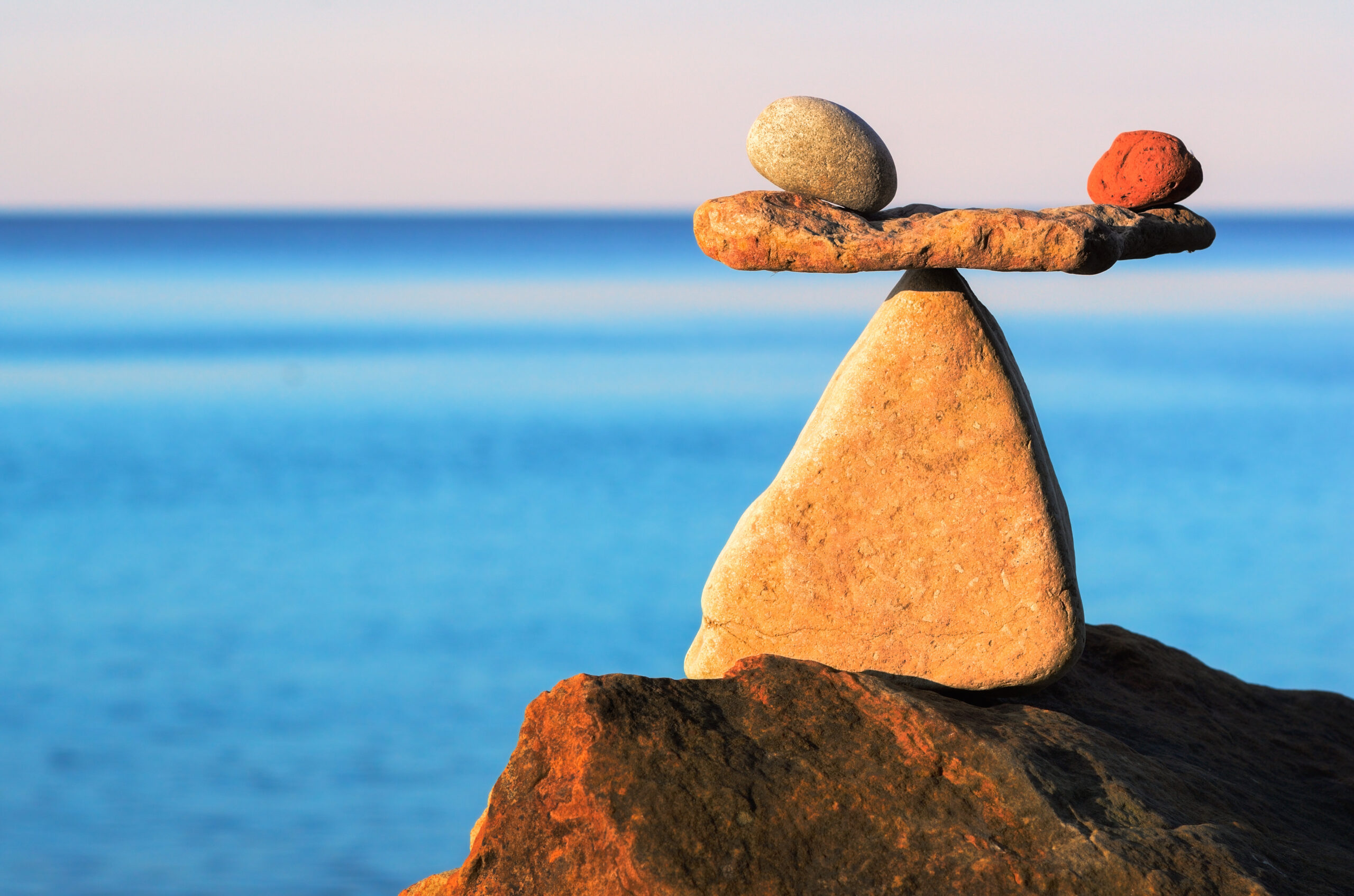 Stacked rocks balanced on a large rock by the blue ocean, with calm water in the background.