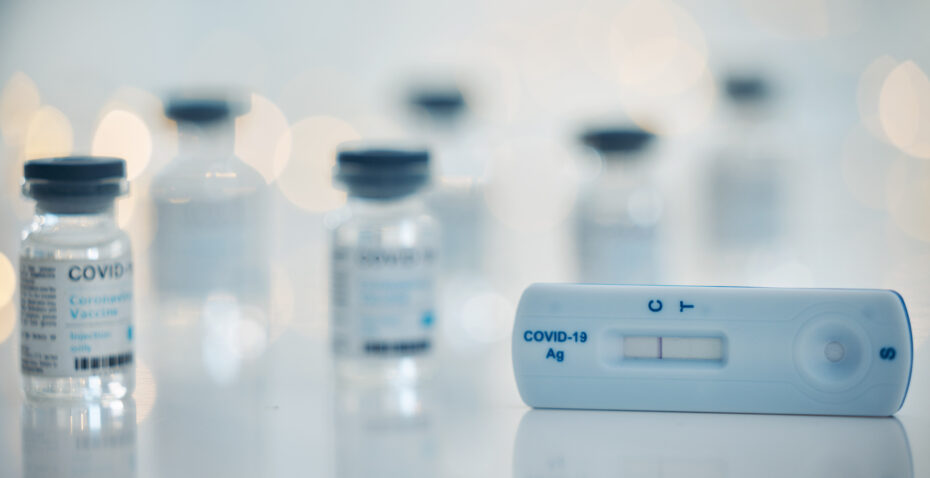 COVID-19 antigen rapid test device on a white surface with several blurred vaccine vials in the background in a clinical setting.