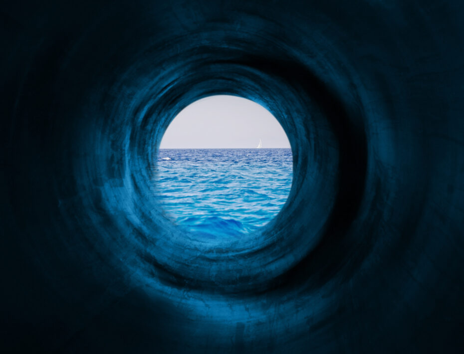 View through a dark circular tunnel opening to a calm blue sea with a small white sailboat on the horizon.