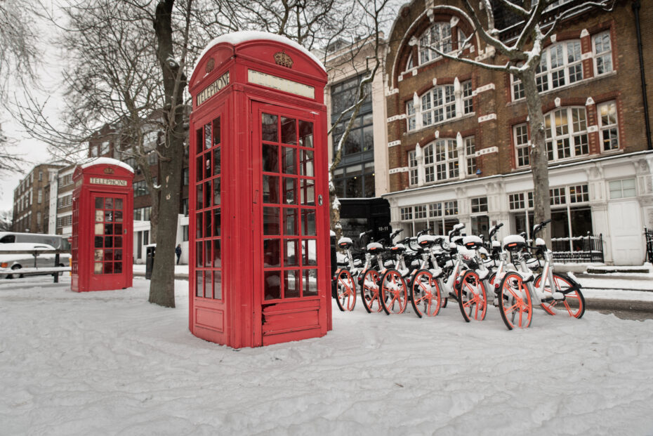 Red classic telephone booth on a snowy city street with a line of orange-red bike-share bicycles in front and brick buildings in the background.