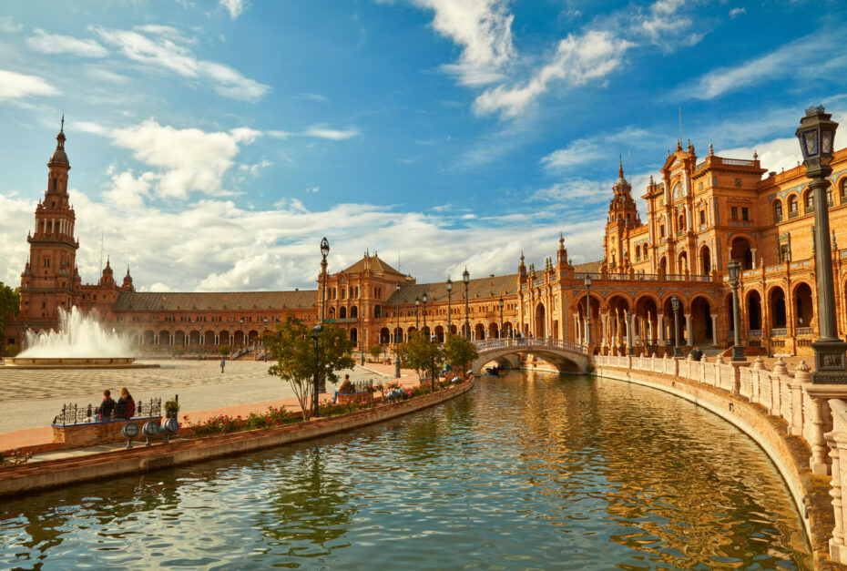Panoramic view of Plaza de España in Seville: red-brick arches, ornate towers, a canal, and a stone bridge under a blue sky.