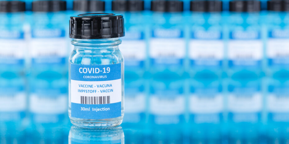 Close-up of a glass vaccine vial labeled COVID-19 in focus, with blurred rows of similar vials in the background on a blue surface.