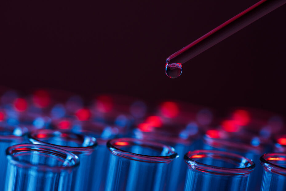 Close-up of a glass pipette releasing a droplet over a row of test tubes in blue and red neon lighting.