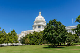 CBO Doesn’t Capture The Real Impact of H.R. 3 on Innovation - OHE White U.S. Capitol dome rises above green lawn and trees under a clear blue sky, with a small flag visible near the building.