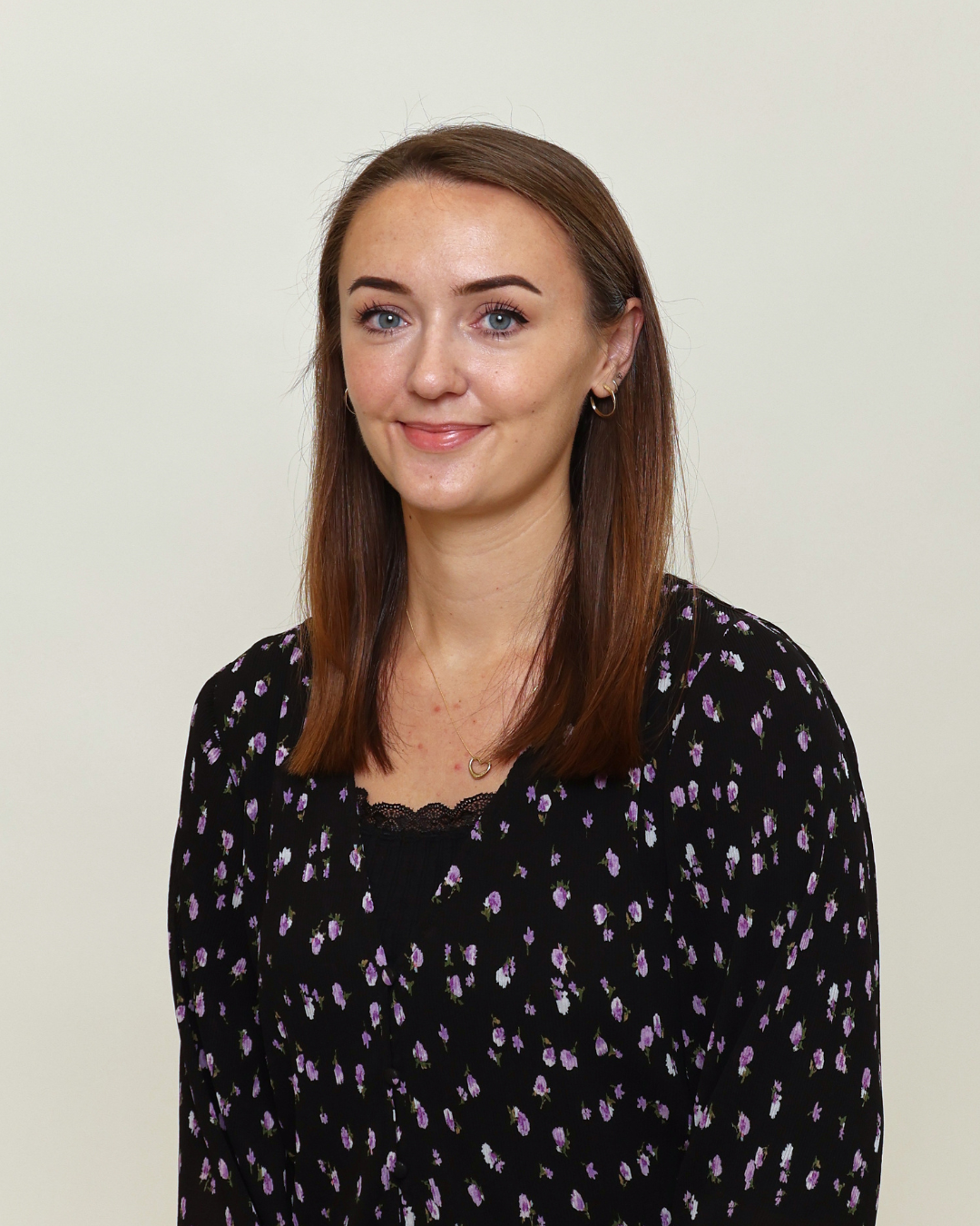 Portrait of a smiling woman with shoulder-length brown hair wearing a black floral blouse and gold hoop earrings against a light background