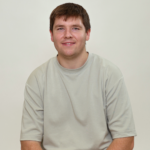 Matthew Napier - OHE Portrait of a smiling man with short brown hair in a light gray T-shirt, seated against a plain beige background.
