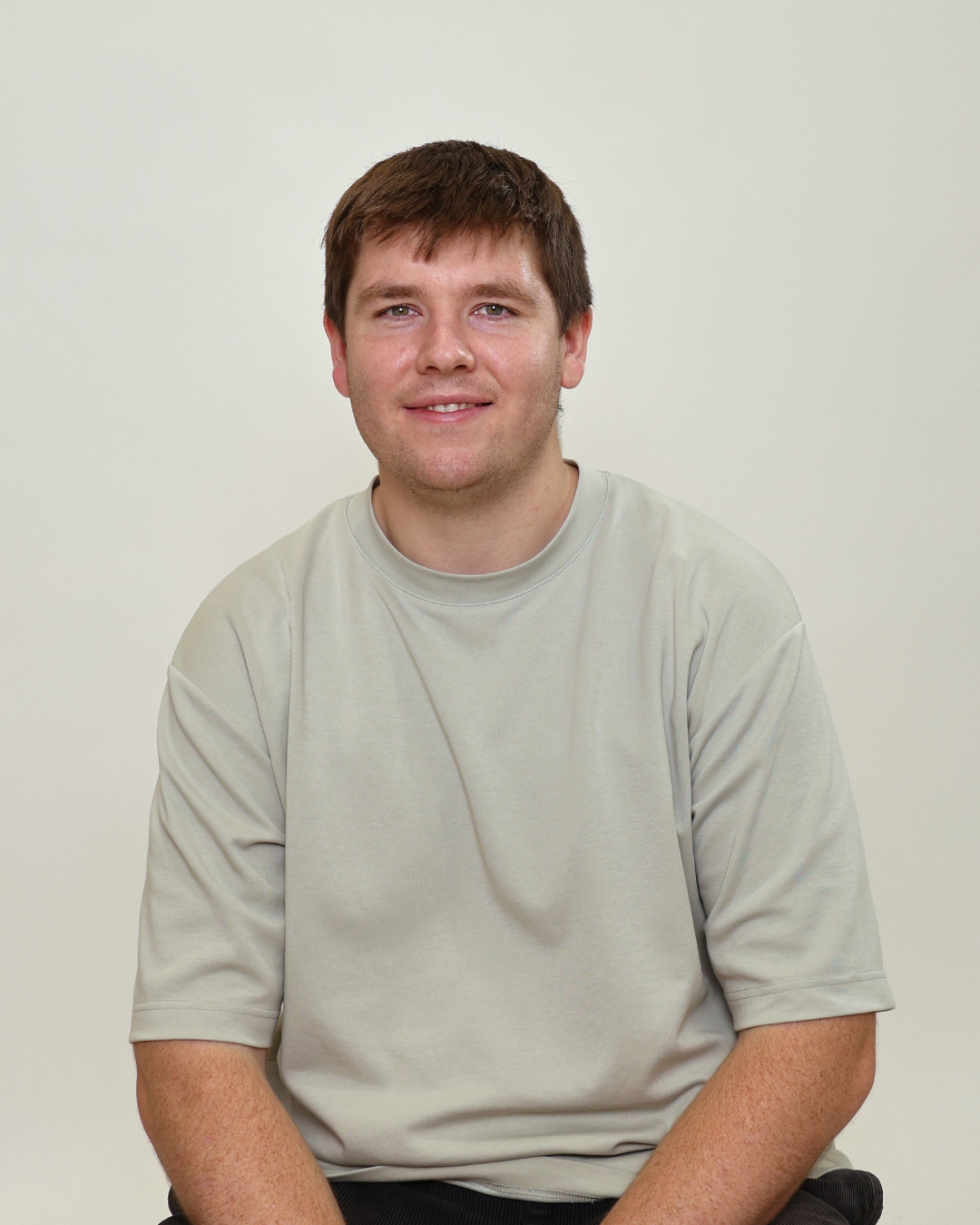 Portrait of a smiling man with short brown hair in a light gray T-shirt, seated against a plain beige background.