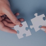 two hands holding two white puzzle pieces close to each other.