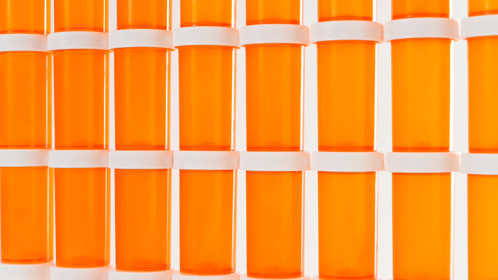 A close-up image of a collection of orange prescription bottles neatly aligned in rows, with white caps and blank labels, against a white background. The repetitive pattern and bright color highlight the pharmaceutical industry’s mass production of medication.