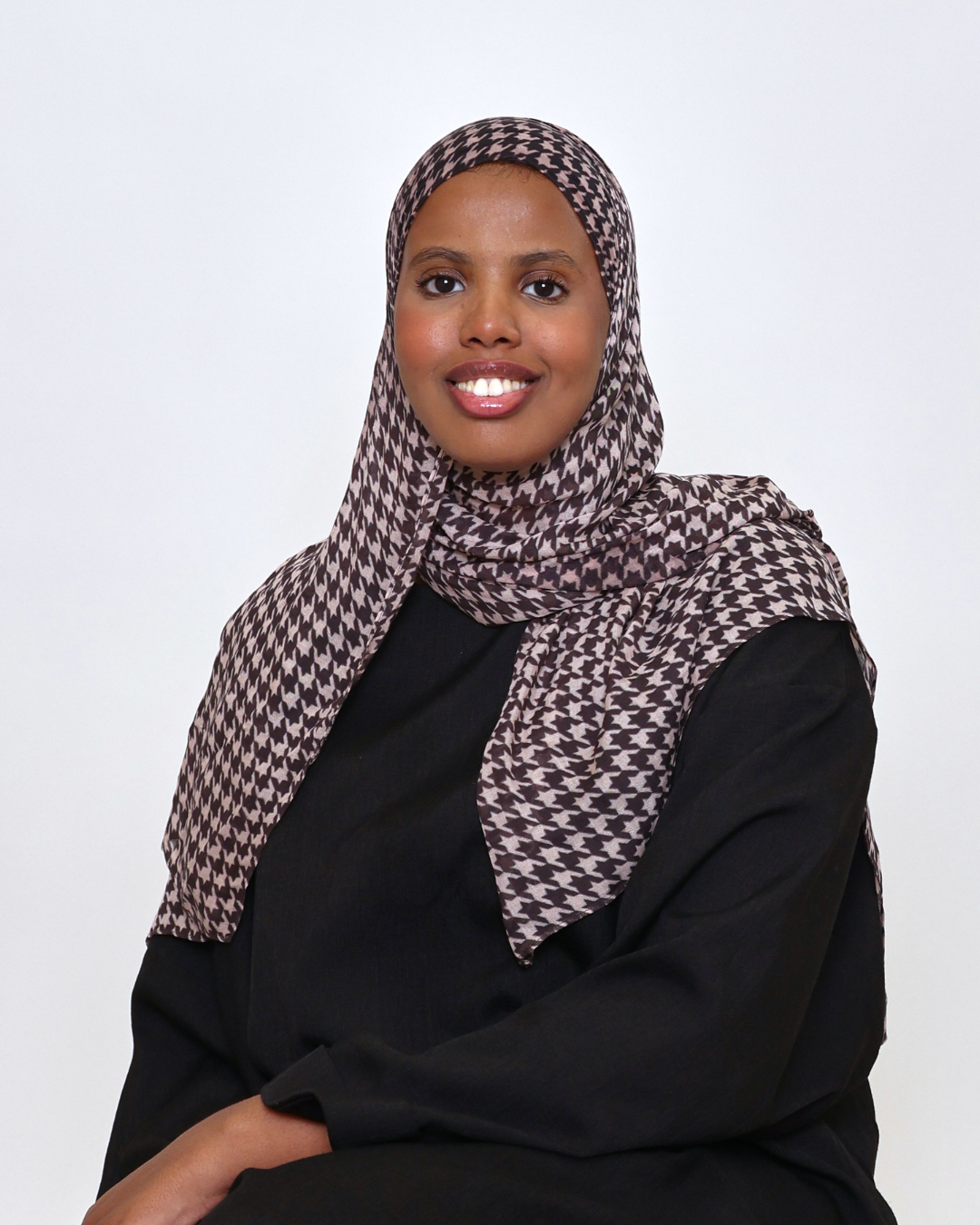 Portrait of a woman wearing a brown and beige patterned hijab and black top, smiling at the camera.