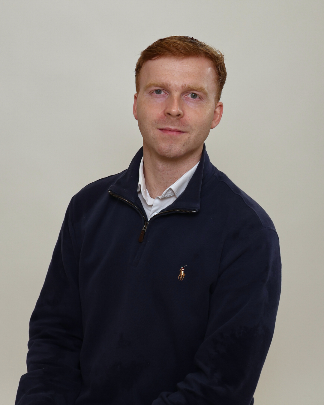 Portrait of a man with short red hair wearing a navy quarter-zip sweater over a white shirt, posed against a light gray background.