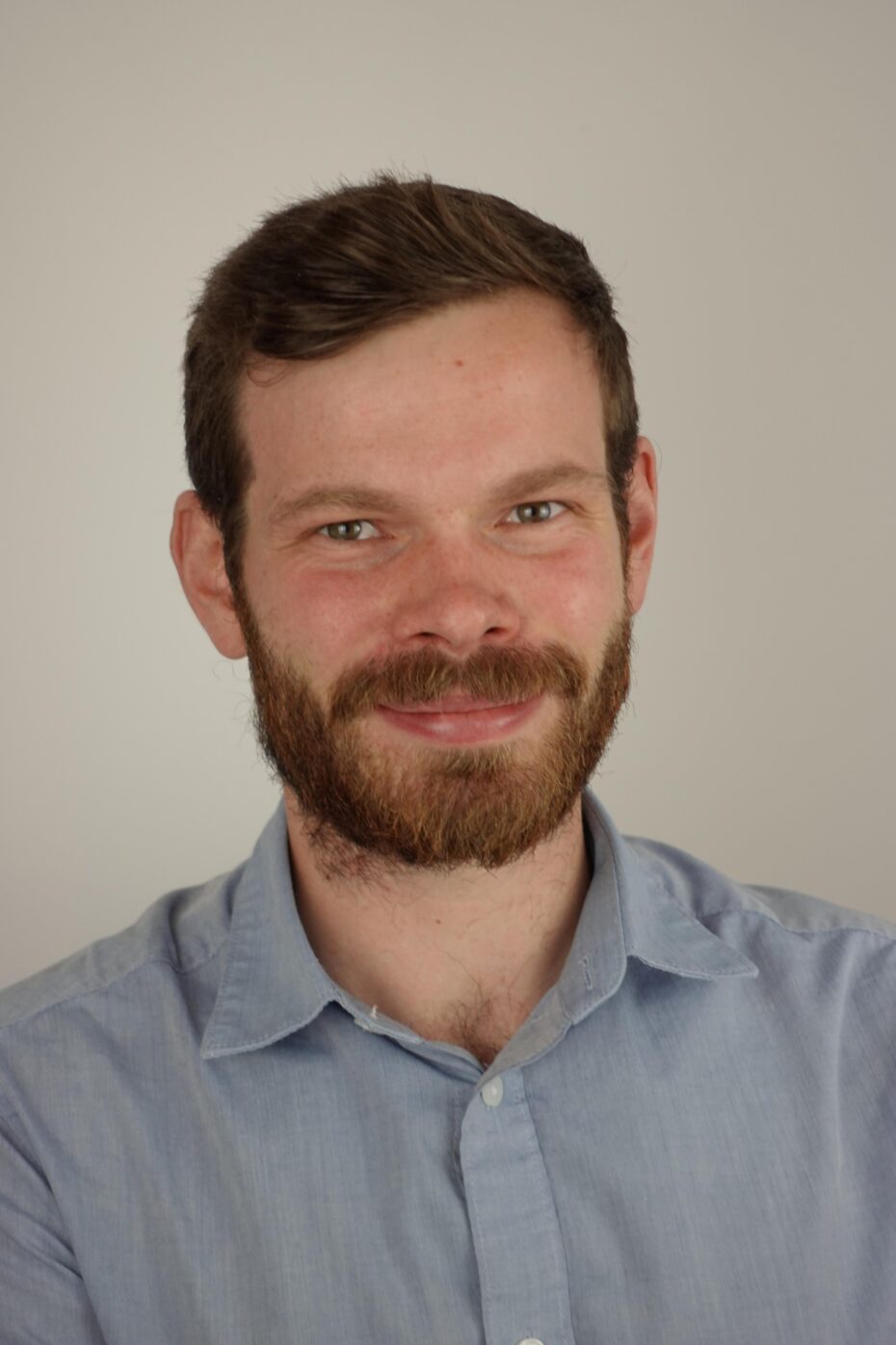 Close-up portrait of a smiling man with short brown hair and a full beard, wearing a light blue shirt against a neutral background.