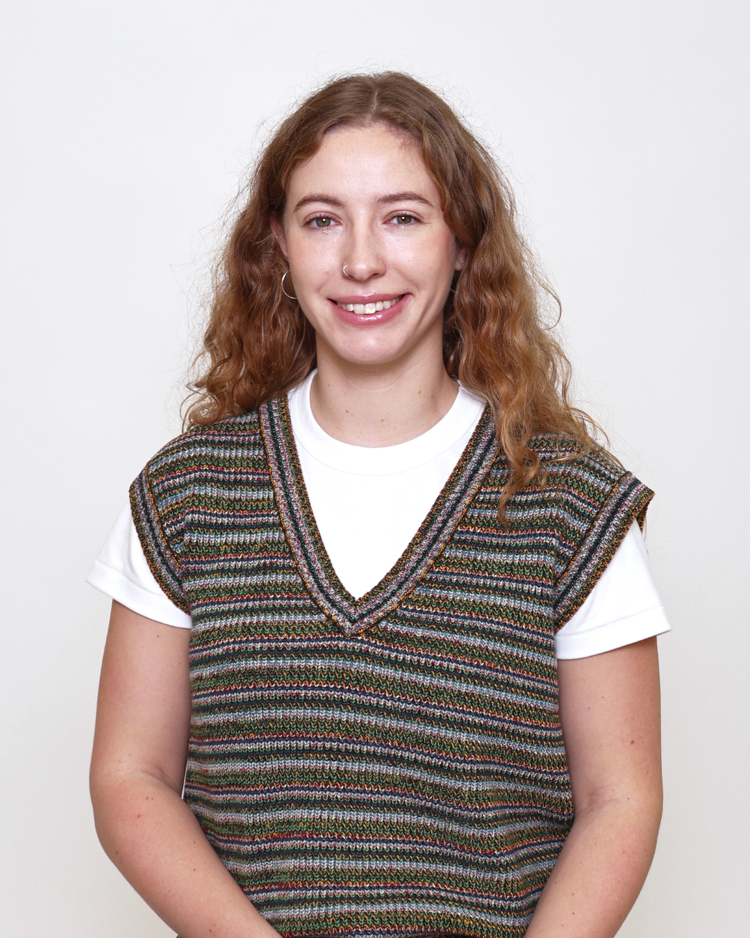 Smiling young woman with curly light brown hair wearing a white T-shirt under a multicolor knit V-neck vest, neutral backdrop.