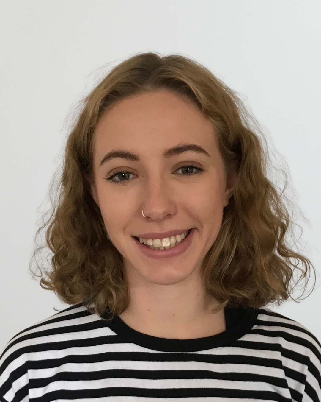 Portrait of a smiling young woman with shoulder-length wavy blond hair wearing a black-and-white striped shirt, facing the camera.