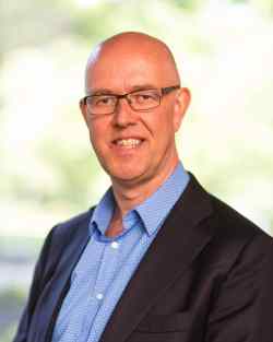 Professional head-and-shoulders portrait of a smiling bald man wearing glasses, a blue shirt, and a dark blazer.