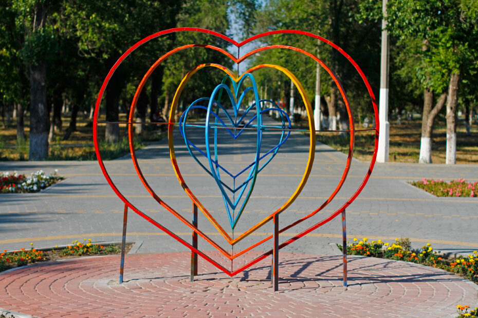 Colorful metal heart sculpture with red, orange, yellow, and blue rings in a tree-lined park on a paved plaza.