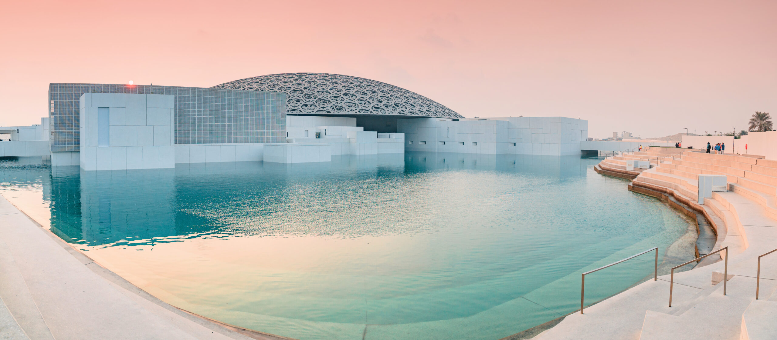 Limestone and glass museum by a reflective turquoise pool at sunset with a latticed dome roof (Louvre Abu Dhabi).