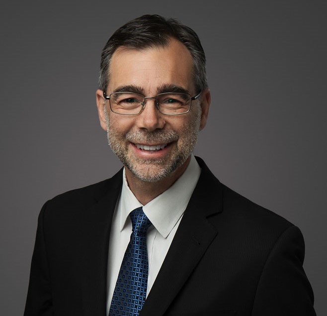 Professional headshot of a smiling middle-aged man with salt-and-pepper hair and glasses, wearing a black suit and blue patterned tie against a gray backdrop.