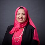 Dalia Dawoud - OHE Smiling woman in pink hijab and pearl necklace, wearing a black blazer against a gray backdrop.