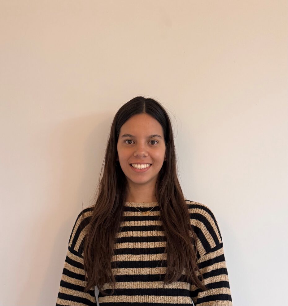 Portrait of a young woman with long brown hair, wearing a black and beige striped sweater, smiling at the camera against a plain beige wall.