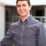 Smiling man in a navy checkered shirt leaning on a white railing indoors.