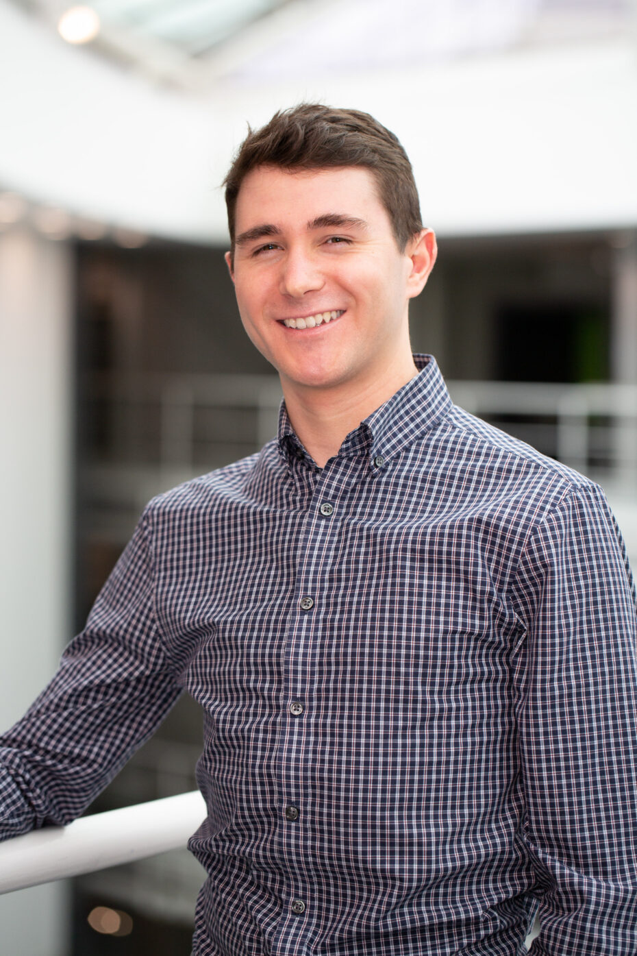 Smiling man in a navy checkered shirt leaning on a white railing indoors.