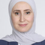 Portrait of a woman in a light gray hijab and navy blazer, smiling softly against a neutral background.