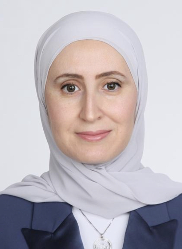 Portrait of a woman in a light gray hijab and navy blazer, smiling softly against a neutral background.