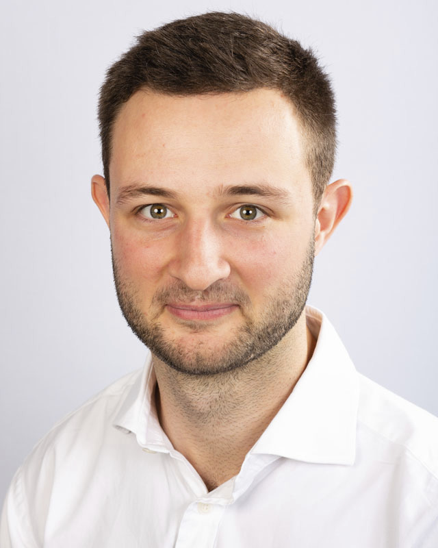 Head-and-shoulders portrait of a man with short dark hair and light stubble, wearing a white shirt and looking at the camera.