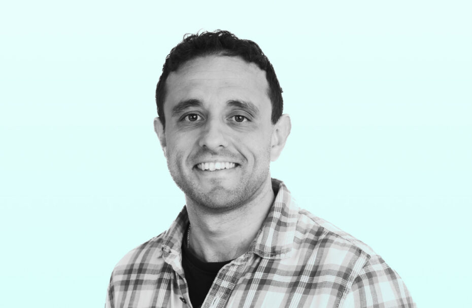 Smiling man in a plaid shirt facing the camera against a light teal background (head and shoulders).