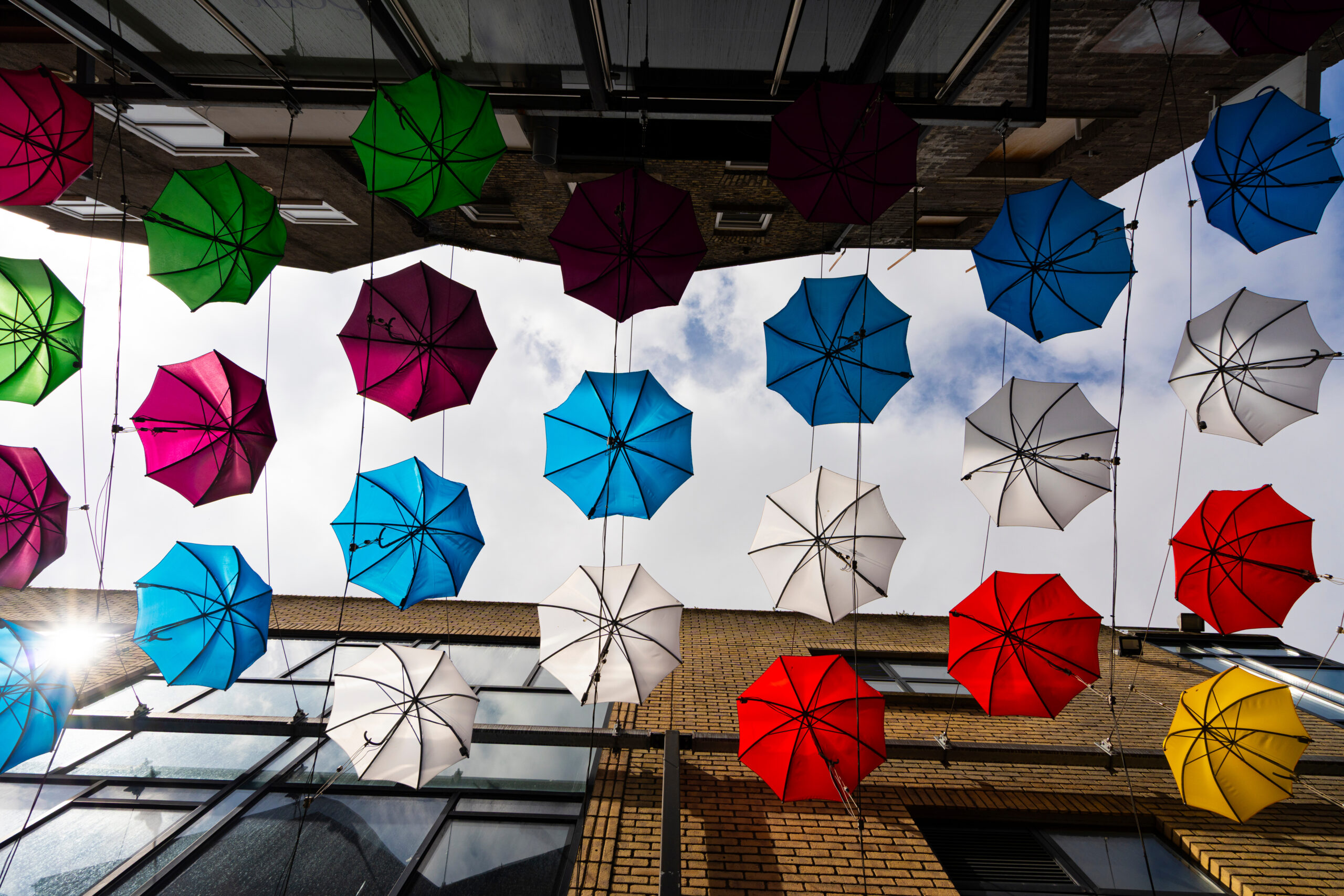 Colorful umbrellas suspended overhead between brick buildings, creating a vibrant canopy above the street.