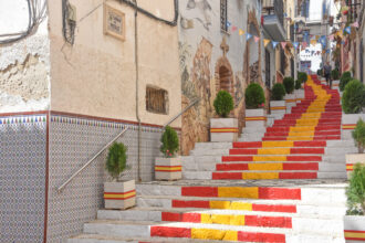 Colorful red and yellow steps descending a narrow street with bunting, murals on the wall, and potted evergreens along the sides.