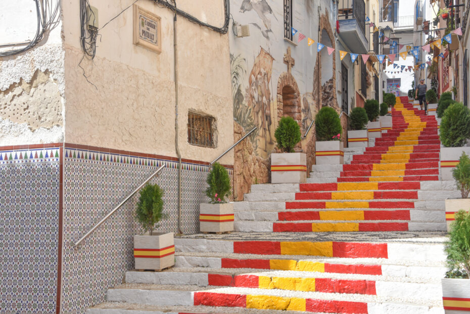 Steps painted in the colour of the Spanish Flag Calpe Alicante Spain - OHE Colorful red and yellow steps descending a narrow street with bunting, murals on the wall, and potted evergreens along the sides.
