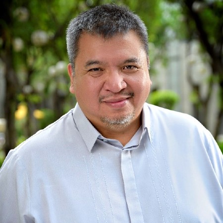 Portrait of a smiling man in a light blue shirt outdoors with greenery in the background.