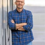 Man with glasses in a blue plaid shirt stands with arms crossed beside a glass wall.