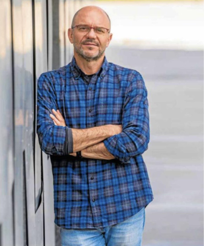 Man with glasses in a blue plaid shirt stands with arms crossed beside a glass wall.