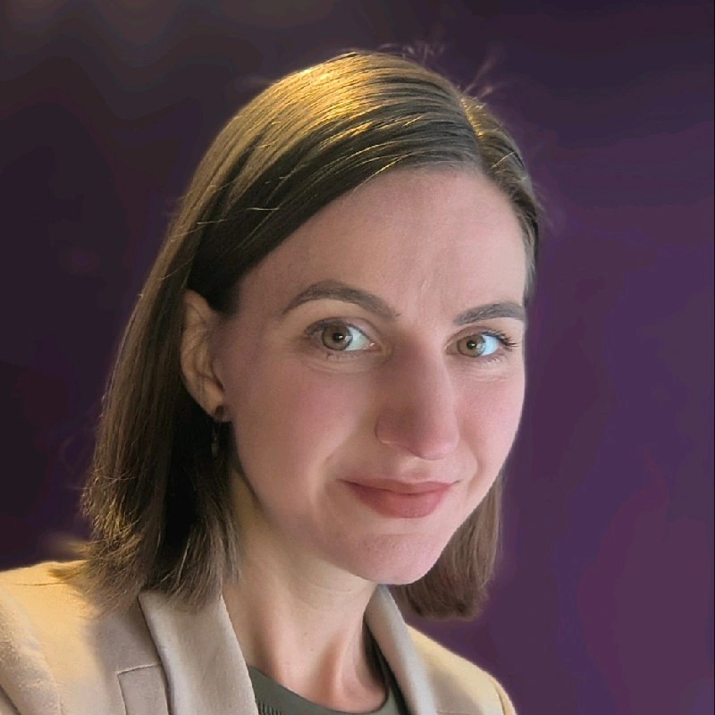 Portrait of a woman with shoulder-length brown hair, wearing a beige blazer, against a purple background.