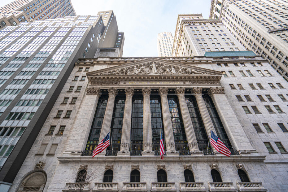 Front facade of the New York Stock Exchange building with classical columns and sculpted pediment, flanked by tall office towers and waving American flags, viewed from street level.