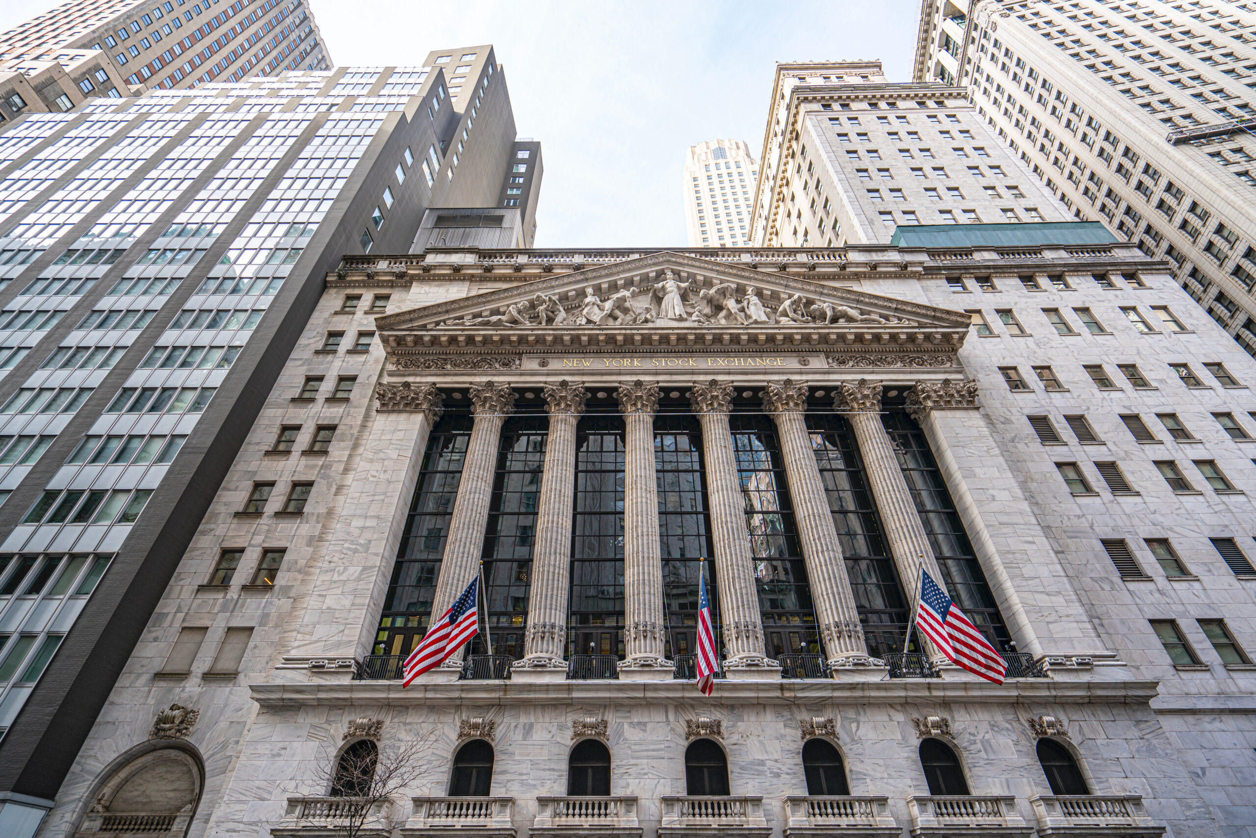 Front facade of the New York Stock Exchange building with classical columns and sculpted pediment, flanked by tall office towers and waving American flags, viewed from street level.