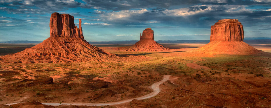 Desert landscape with three massive red sandstone buttes rising from a rocky plain under a blue sky with scattered clouds, a dirt road winding toward them at center.