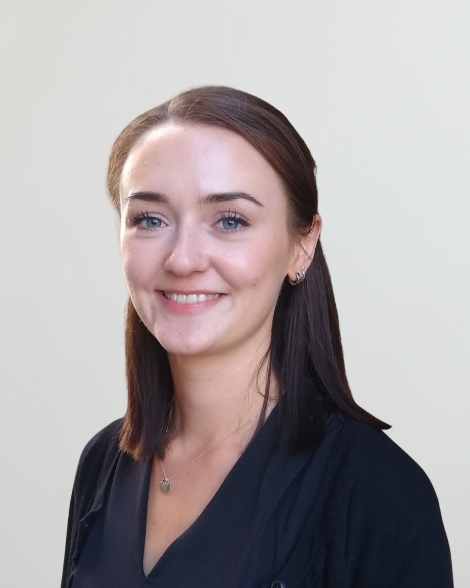 Portrait of a smiling woman with dark hair and blue eyes, wearing a black top and a heart pendant necklace, neutral beige background.