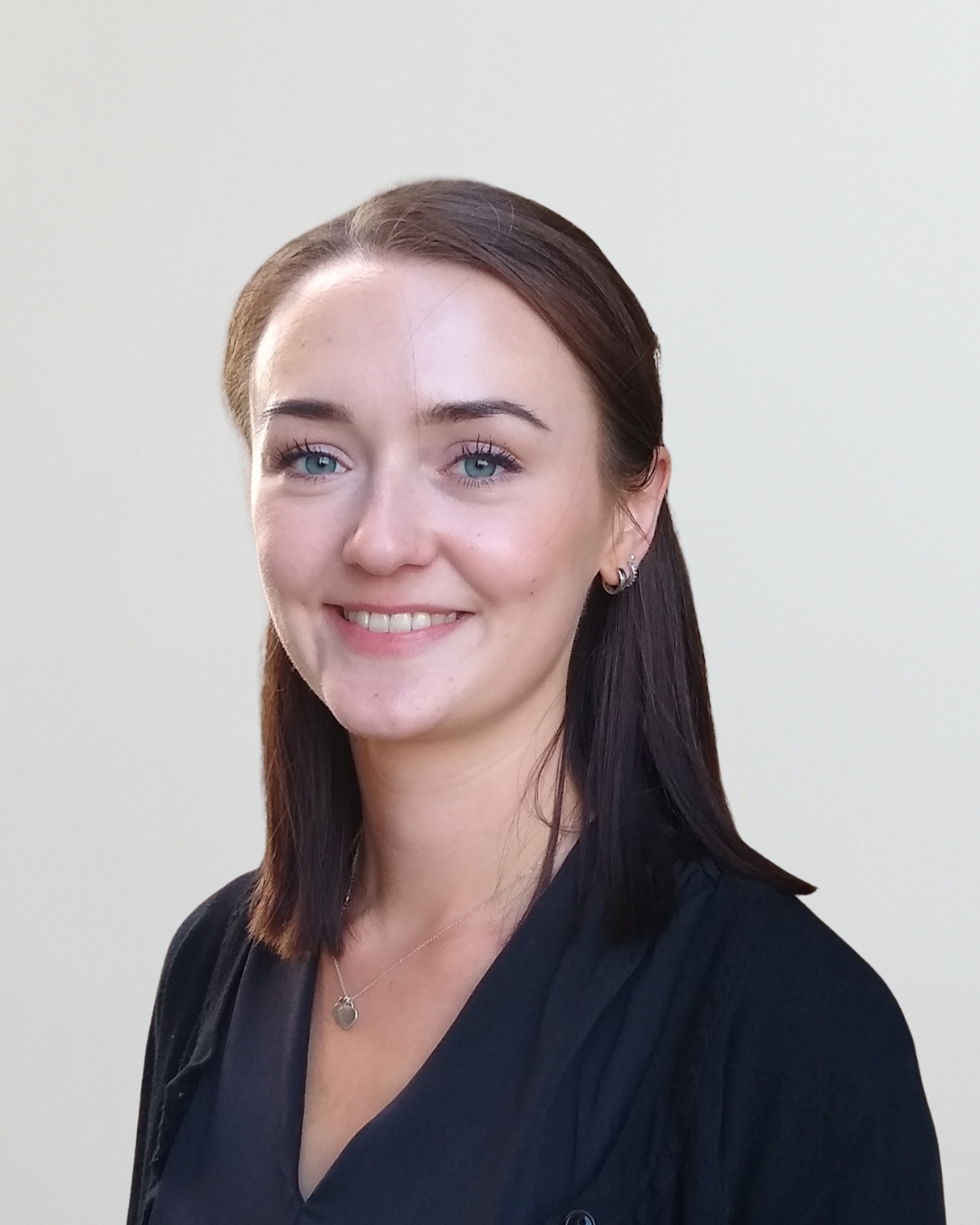 Portrait of a smiling woman with dark hair and blue eyes, wearing a black top and a heart pendant necklace, neutral beige background.