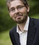 IMG_1426 - OHE Portrait of a bearded man wearing glasses, a dark blazer and white shirt, outdoors with greenery in the background.
