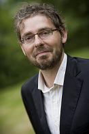 Portrait of a bearded man wearing glasses, a dark blazer and white shirt, outdoors with greenery in the background.