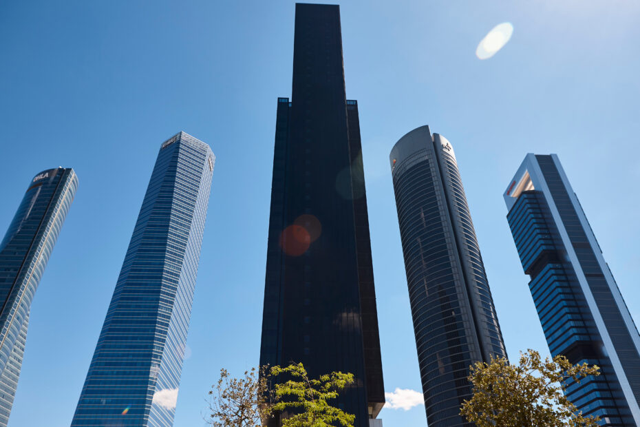 Low-angle view of tall glass skyscrapers reaching up into a clear blue sky with a bit of greenery at the bottom edge.
