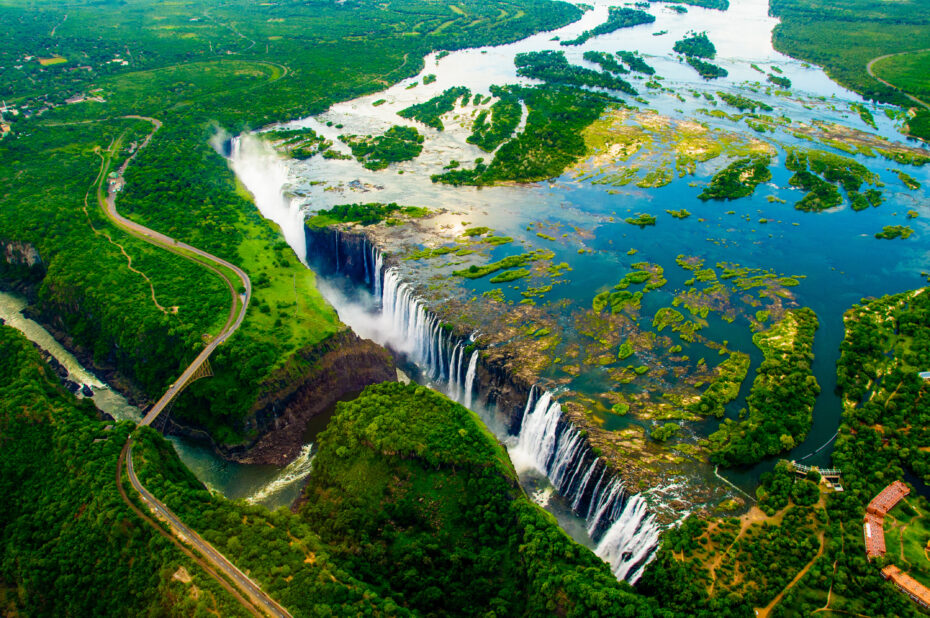 Victoria Falls - OHE Aerial view of a large waterfall cascading into a winding river surrounded by dense green rainforest and bridges nearby.